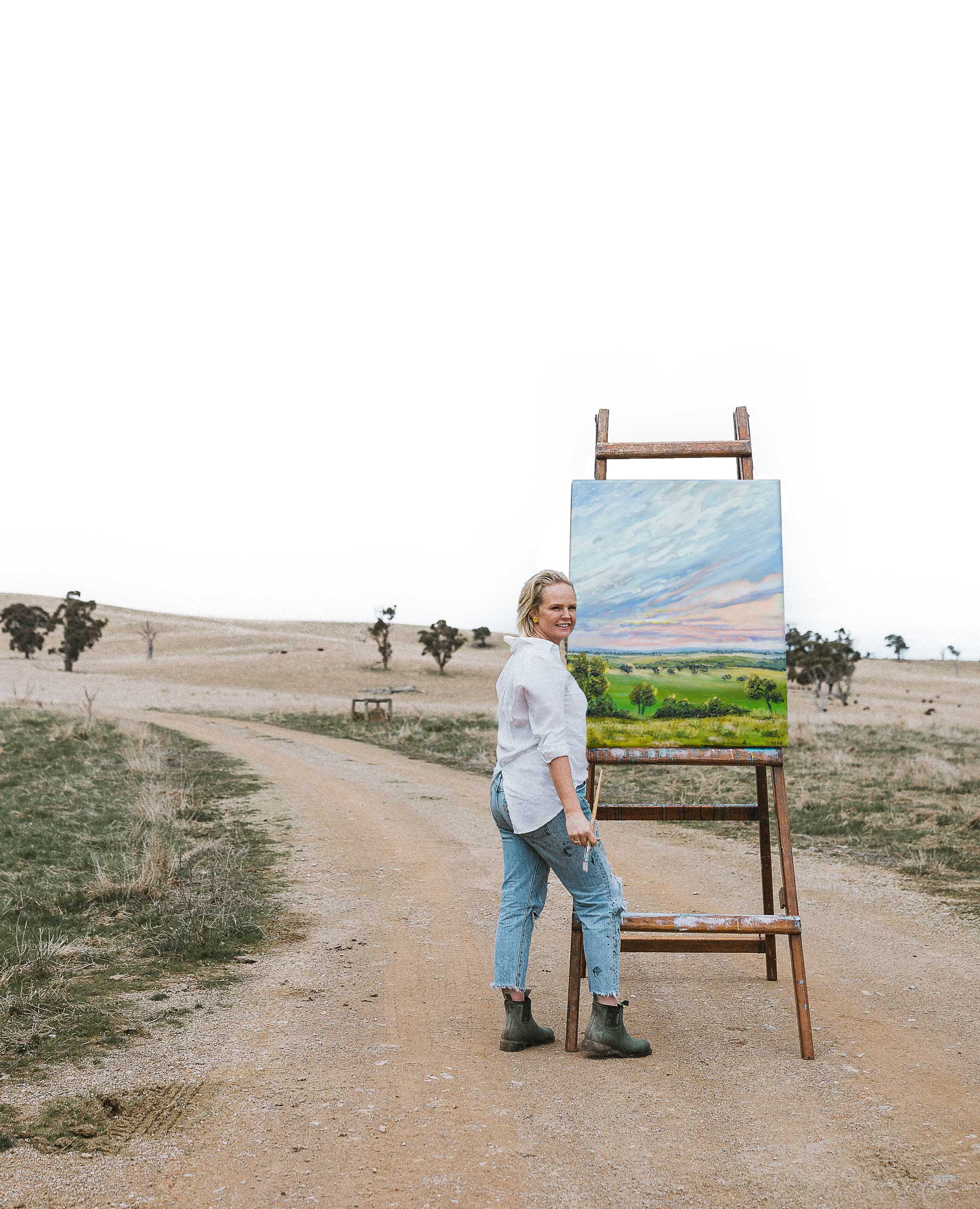 Australian artist, Carina Chambers stands on a dirt road in a rural landscape, leaning against a wooden easel holding a colorful landscape painting. She is wearing a white button-up shirt, light blue jeans, and dark boots.